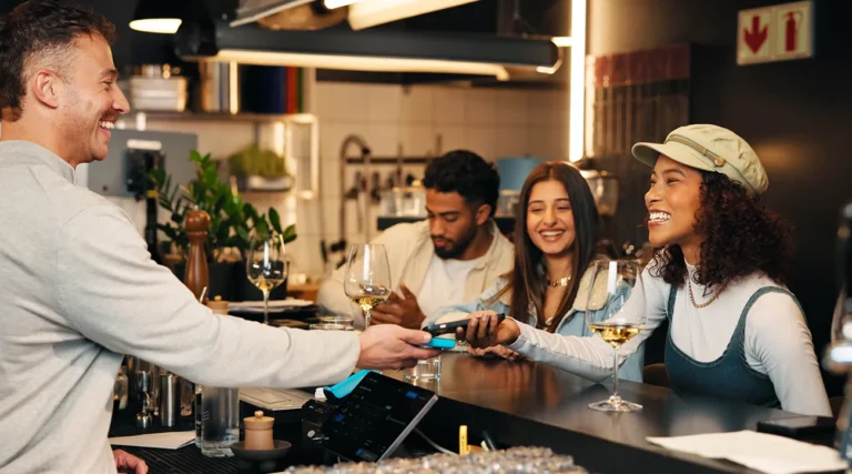 Customer making a contactless card payment at a bar while enjoying drinks with friends