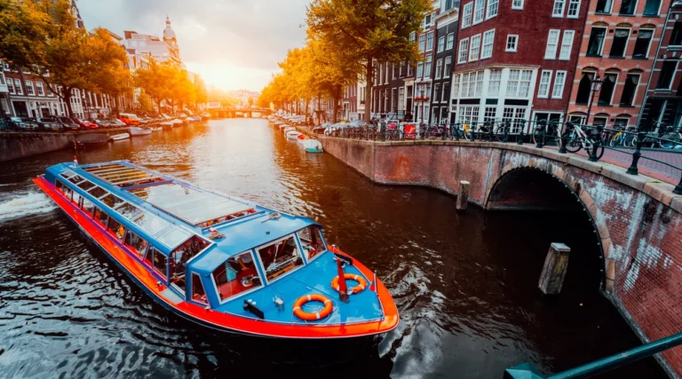 A sightseeing boat cruising past a brick bridge and canal houses in Amsterdam at sunset