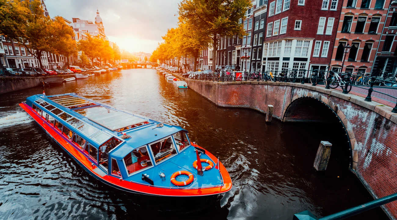 A sightseeing boat cruising past a brick bridge and canal houses in Amsterdam at sunset