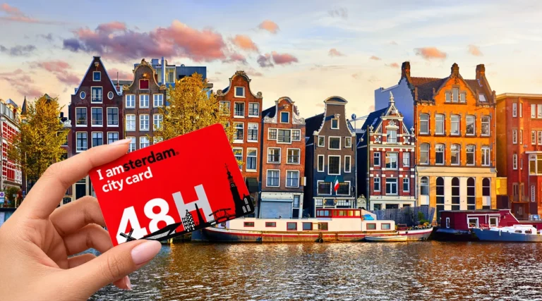 Female hand holding a red I amsterdam City Card 48H, with traditional Dutch canal houses and boats along an Amsterdam canal