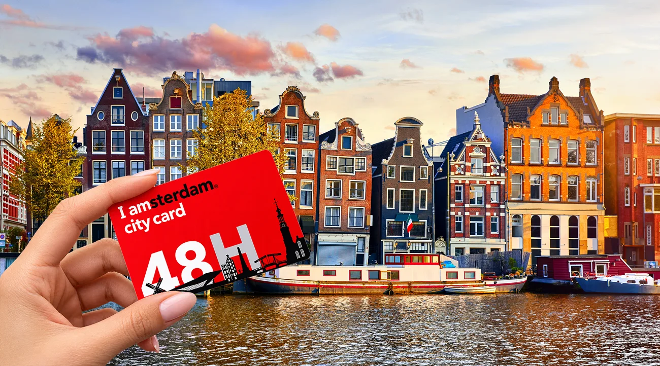 Female hand holding a red I amsterdam City Card 48H, with traditional Dutch canal houses and boats along an Amsterdam canal
