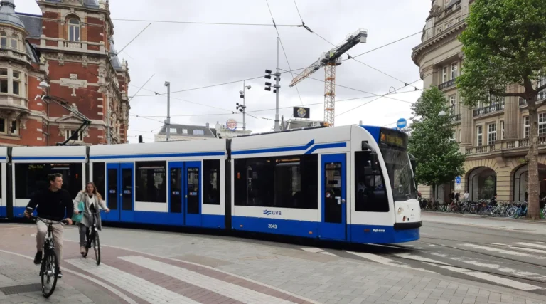 Blue and white GVB tram crossing a central Amsterdam square beside cyclists and historic buildings
