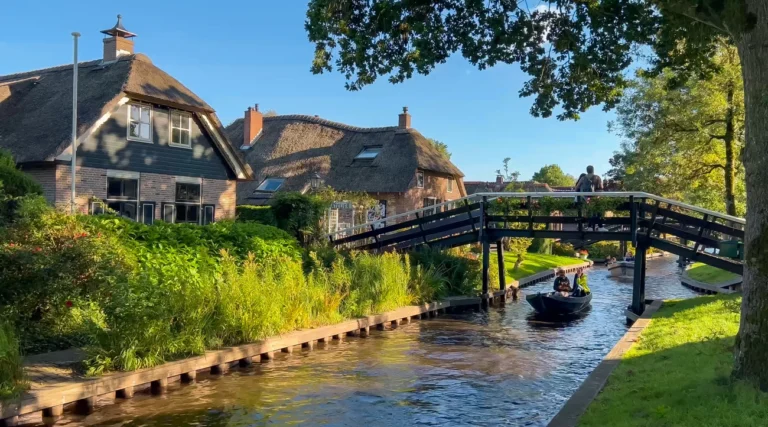 Boat passing beneath a wooden footbridge beside thatched-roof houses in Giethoorn on a sunny day