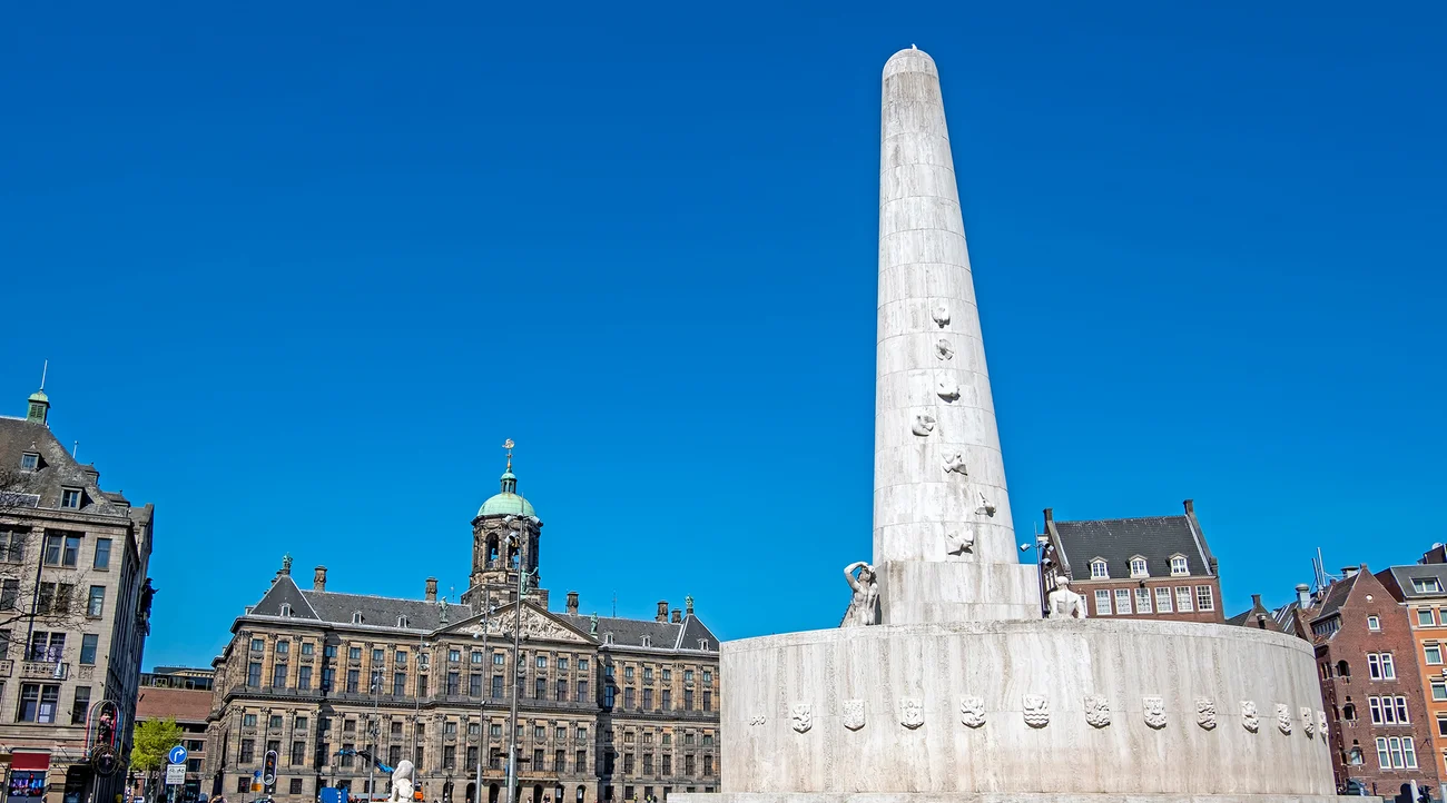 National Monument on Dam Square in Amsterdam beneath a clear blue daytime sky, with the Royal Palace behind