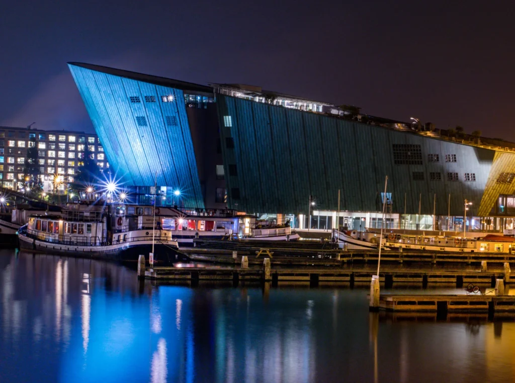 Closer night view of NEMO Science Museum illuminated in blue, with boats and harbour reflections in Amsterdam